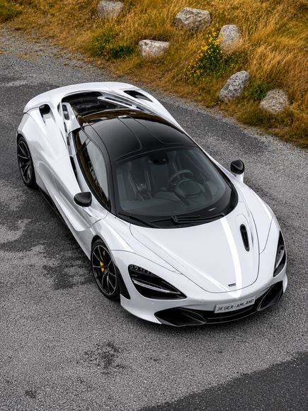 White sports car parked on a roadside, surrounded by green grass, yellow flowers, and rocks, viewed from above by drone.