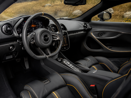 Interior angle of a car, showing black leather seats, steering wheel, speedometer, and buttons, with grass and rocks visible through the window.