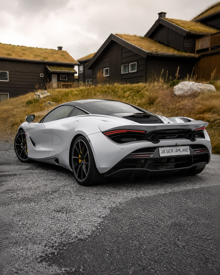 White sports car, rear three-quarter view, parked on grass near a house, with clear sky in the background.