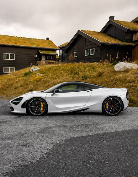 White sports car in side-profile, parked before a house with a green roof, surrounded by grass and rocks, with clear sky in background.
