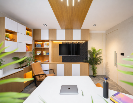A left-side view of a modern office with a white table, laptop, books, and plants. A television is mounted on the wall, and the ceiling has lights.