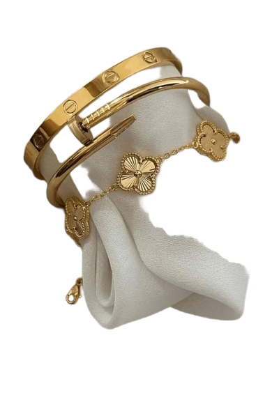 Two gold bangles and a clover-motif bracelet resting on draped white silk — background removed by Photoroom