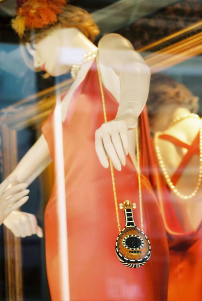 Shop mannequin in an orange dress with a pendant necklace, seen through glass — upscaled by GoStudio Lite