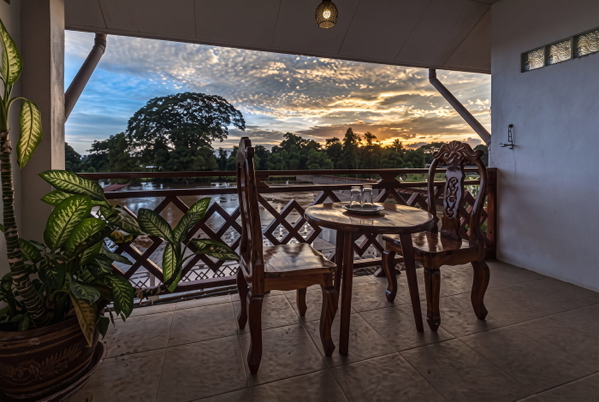 Balcony with carved wooden chairs and a table overlooking a river at sunset — upscaled by GoStudio Advanced