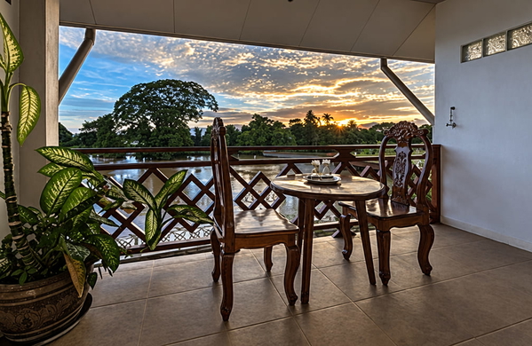 Balcony with carved wooden chairs and a table overlooking a river at sunset — upscaled by GPT Image 2.5