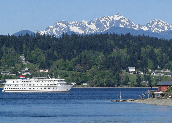 Small white cruise ship on blue water below forested hills and snow-capped mountains — watermark removed by GoStudio Advanced