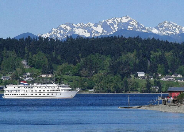 Small white cruise ship on blue water below forested hills and snow-capped mountains — watermark removed by GoStudio Lite