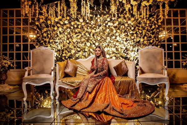 Bride in an orange embroidered lehenga seated on a stage of gold flowers and chairs — watermark removed by GoStudio Advanced