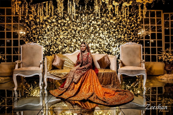 Bride in an orange embroidered lehenga seated on a stage of gold flowers and chairs