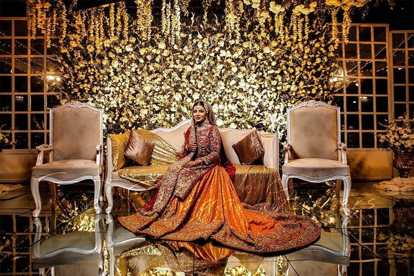 Bride in an orange embroidered lehenga seated on a stage of gold flowers and chairs — watermark removed by GoStudio Lite