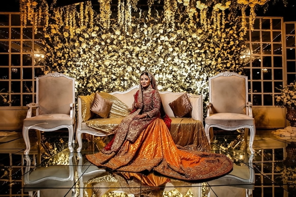 Bride in an orange embroidered lehenga seated on a stage of gold flowers and chairs — watermark removed by GoStudio Pro