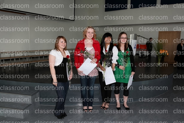Four graduates holding certificates and flowers on a hall stage