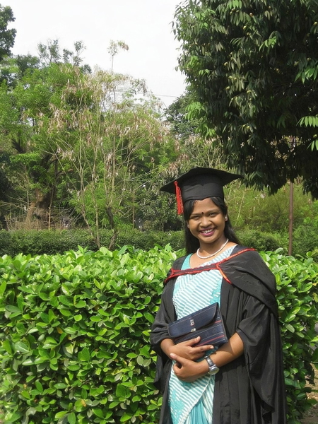 Graduate in a black gown and cap over a pale blue dress, in front of a hedge — watermark removed by GoStudio Advanced