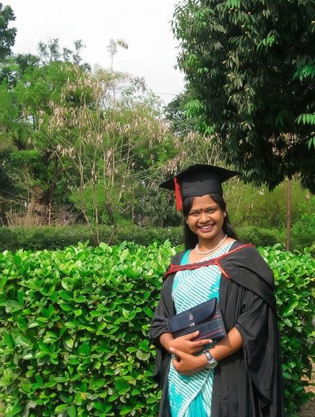 Graduate in a black gown and cap over a pale blue dress, in front of a hedge — watermark removed by GoStudio Pro