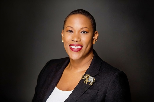 Woman in a navy blazer with an elephant brooch, smiling, on a dark backdrop — watermark removed by GoStudio Advanced