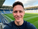 A smiling man in a t-shirt taking a selfie by a soccer field — headshot before AI full-body generation.
