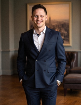 A man in a navy suit smiles for a professional headshot, standing in front of a painting. A man in a navy suit smiles for a professional headshot, standing in front of a painting.