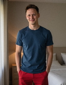 A man smiling, wearing a navy blue t-shirt and red pants, standing in a bedroom before AI virtual try-on. A man smiling, wearing a navy blue t-shirt and red pants, standing in a bedroom before AI virtual try-on.