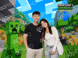 A man and woman smiling in front of a Minecraft-themed backdrop of colorful blocks, shown with a watermark on the photo.