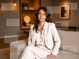 A woman in a white dress sitting on a couch in a living room with wall art, bookshelves and a plant, shown with a watermark.