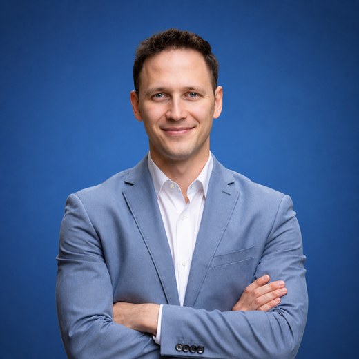 The image shows a man in a blue suit with his arms crossed, smiling against a blue background. The image shows a man in a blue suit with his arms crossed, smiling against a blue background.