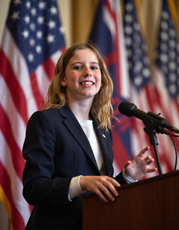 A woman smiles at a podium with an American flag, holding a microphone.