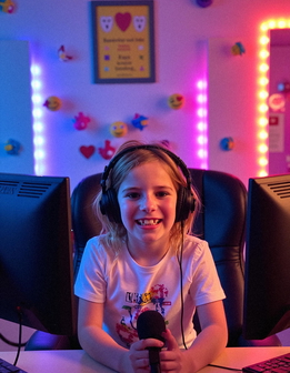 A child sits at a desk with two monitors, wearing headphones and holding a microphone.