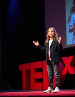 A young person stands on a stage, arms outstretched, wearing a black blazer, white shirt, and jeans.