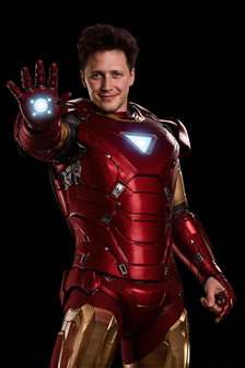 A man in a red and gold suit with a glowing hand, smiles against a black background.