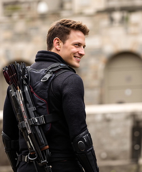 A man in a black jacket, holding a bow and arrow, smiles in front of a building.