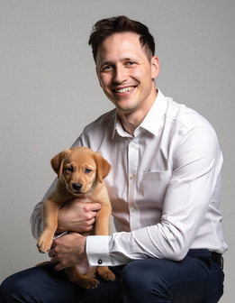 A man in a white shirt and blue pants sits on a stool, smiling while holding a puppy. A man in a white shirt and blue pants sits on a stool, smiling while holding a puppy.