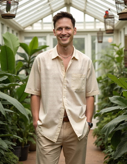 A man smiles in a greenhouse, surrounded by lush plants and bird cages hanging from the ceiling. A man smiles in a greenhouse, surrounded by lush plants and bird cages hanging from the ceiling.