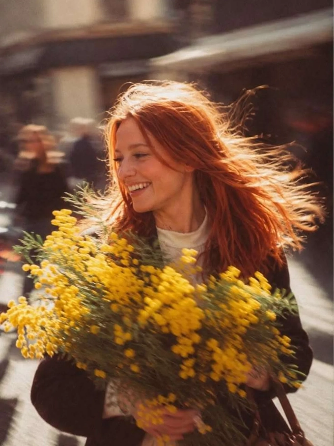 A smiling red-haired woman carrying flowers, her hair and the street in motion while her face stays sharp. A smiling red-haired woman carrying flowers, her hair and the street in motion while her face stays sharp.